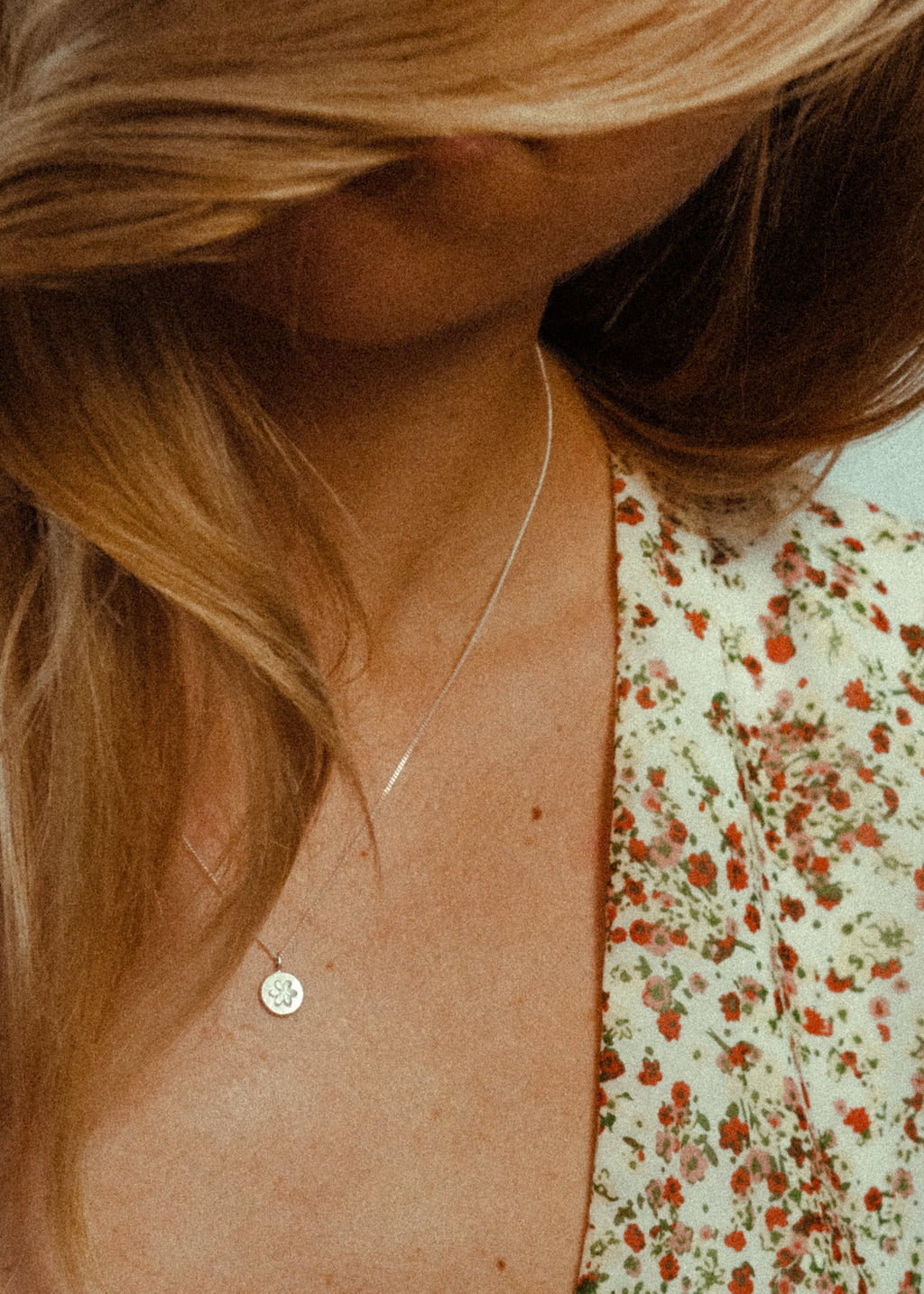 Close-up of a person wearing a delicate necklace with a small round pendant, against a blurred floral-patterned background.