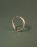 A minimalist hazelnut gold court band ring standing upright on a dark green textured background.