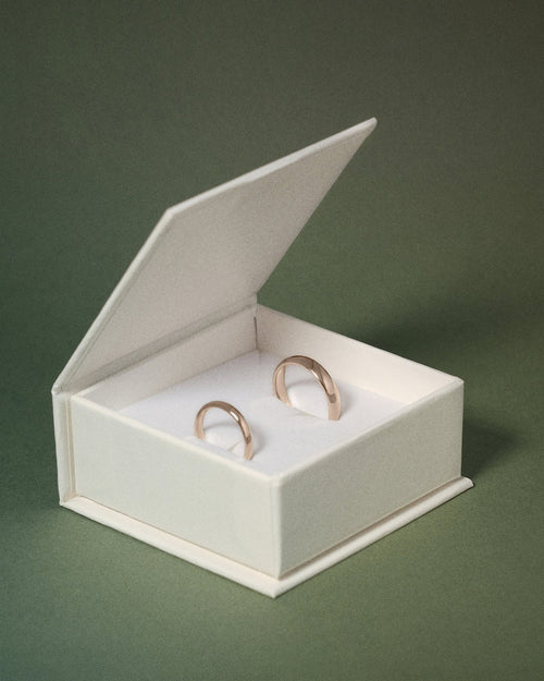 Two slim hazelnut gold wedding rings displayed inside an open white jewelry box on a dark green background.