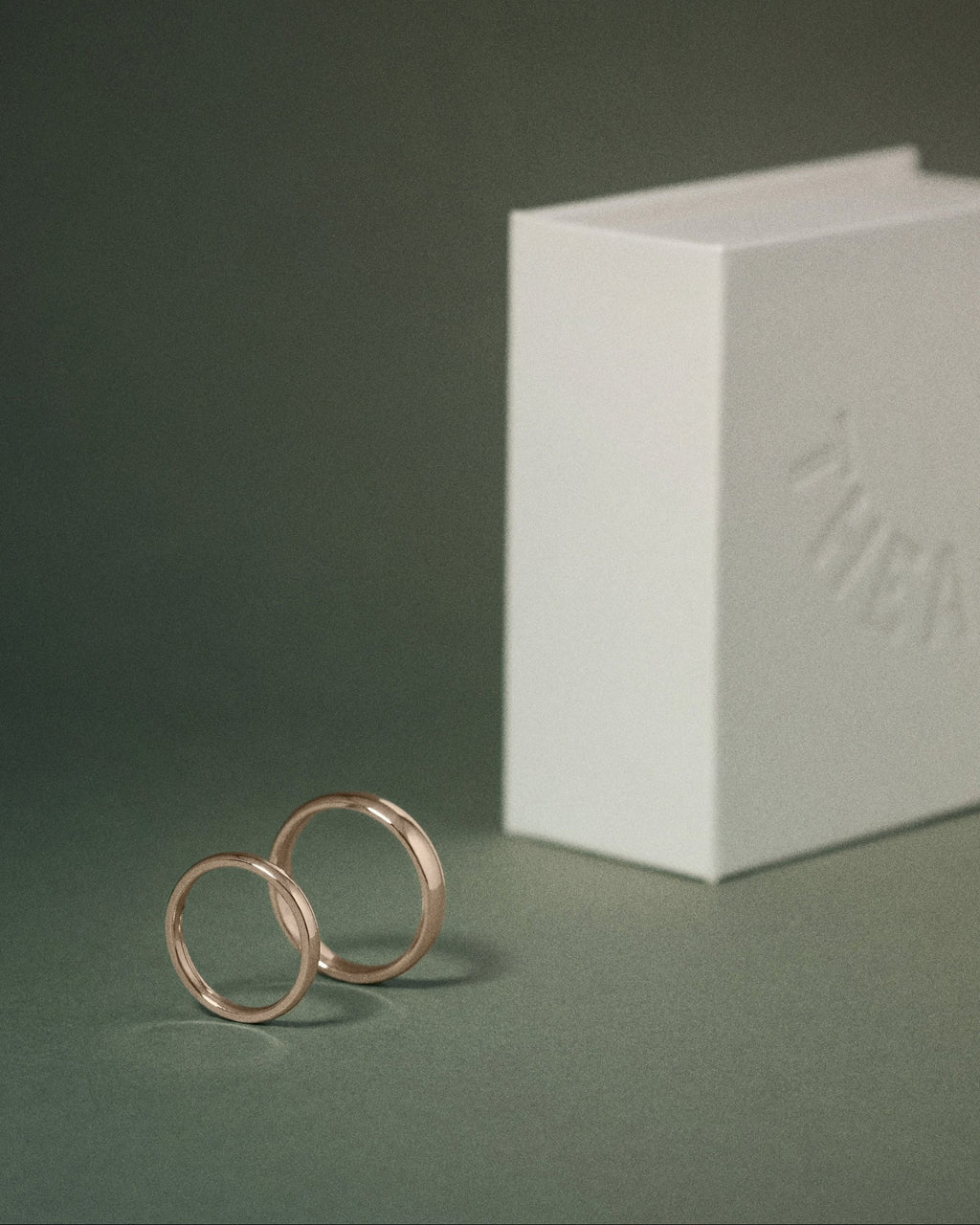 Two slim hazelnut gold court rings standing upright next to a white embossed THEA box on a dark green background.