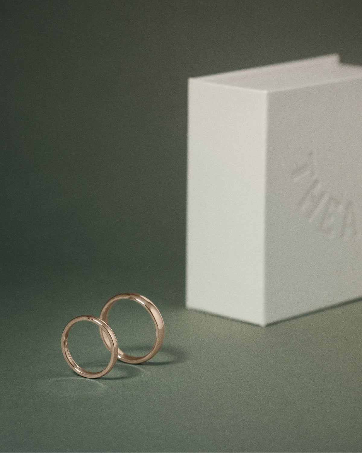 Two slim hazelnut gold court rings standing upright next to a white embossed THEA box on a dark green background.