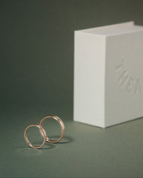Two slim hazelnut gold court rings standing upright next to a white embossed THEA box on a dark green background.